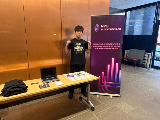 A young man stands at a table holding up a t-shirt that reads 'build the future here', next to a banner for 'NYU BLOCKCHAIN LAB' which states 'CONNECTING STUDENTS WITH THE BLOCKCHAIN INDUSTRY TO UNLOCK REAL-WORLD OPPORTUNITIES'.