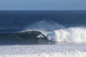 A surfer rides a large, powerful wave with white spray crashing around them in the deep blue ocean.