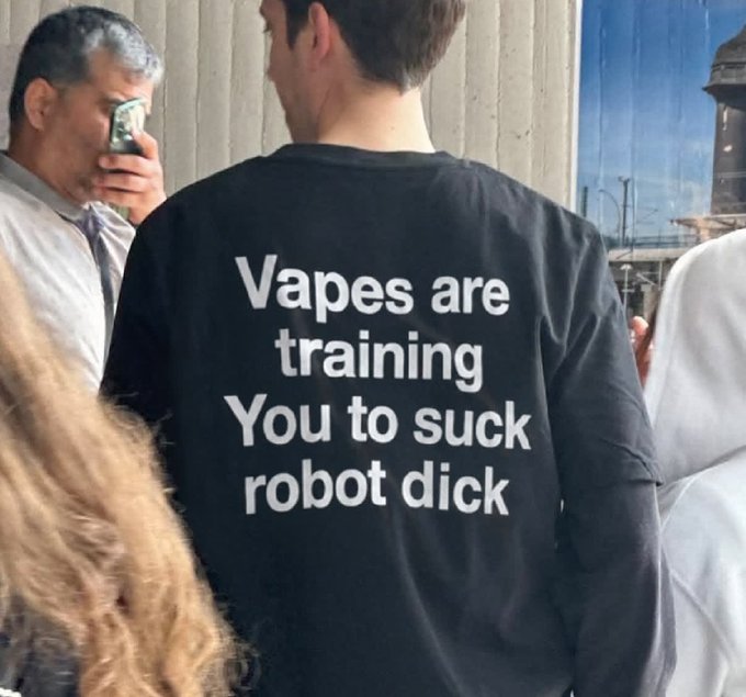 A man wears a black T-shirt reading, “Vapes are training You to suck robot dick.” The accompanying post says “Think about it,” framing the provocative slogan as a commentary on vaping’s gradual normalization and addictive design.
