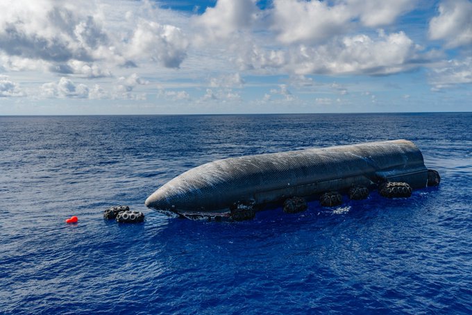 A large cylindrical structure, possibly a submarine or industrial vessel, lies partially submerged in the open ocean under a partly cloudy sky. Several small inflatable boats are clustered around its sides, with one towing a red buoy, suggesting an active operation or investigation—fitting for a post claiming this dramatic maritime scene is "Not AI."