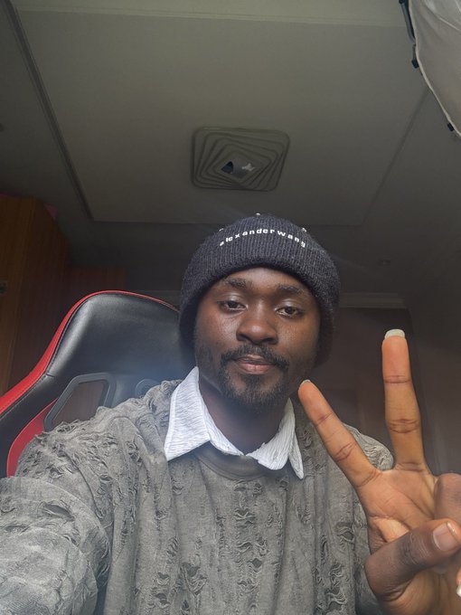 Selfie of a man wearing a gray beanie and a distressed gray sweater over a collared shirt, holding up a peace sign while sitting in a gaming chair.