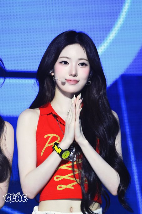 A young woman with long dark hair performing on stage, wearing a red sleeveless top with yellow lettering and a headset microphone, with hands pressed together. The watermark 'GL:G' appears in the corner.