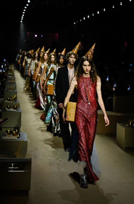 Models walk down a runway wearing sequined dresses and party hats during a fashion show.