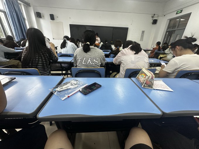 A classroom scene viewed from a student's desk shows classmates seated at blue desks facing a chalkboard, with a smartphone and pens resting on the desk.