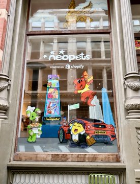 A storefront window display features "Neopets" branding alongside "Toys"R"Us shopify" text, showcasing plush toys and a decorated car with the Neopets character imagery. The image visually represents the Neopets brand itself, the subject of the post's commentary on the company's failed NFT venture and subsequent corporate pivot.