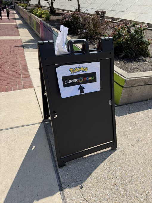 A black A-frame sidewalk sign on a paved walkway displays a printed paper reading 'Pokémon SUPER NOVA' with an upward-pointing arrow.