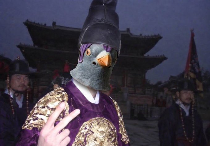A person wearing a grey pigeon head mask and a purple and gold traditional Korean royal robe, posing with a peace sign in front of a palace at dusk.