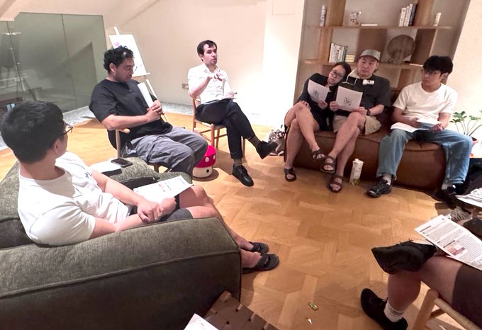 A group of people sitting in a casual living room setting, appearing to be in a discussion or meeting, with some holding papers and pamphlets.