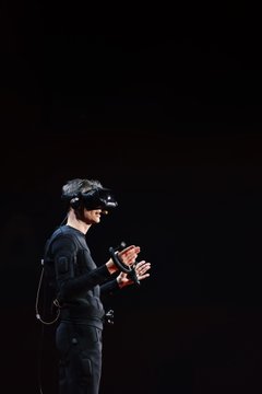 A person in a black motion capture suit wearing a VR/AR headset and holding controllers, standing against a dark background.