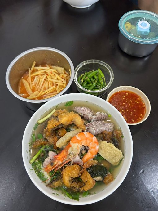 A bowl of Vietnamese soup with shrimp, meat, and fried items is served alongside side dishes of shredded vegetables, herbs, and chili sauce.