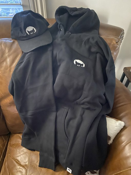 A black Gutter Cat Gang hoodie and matching cap featuring the iconic white cat logo are displayed on a leather couch, representing the merch prize from the Gutter Cat Gang giveaway won by the user.