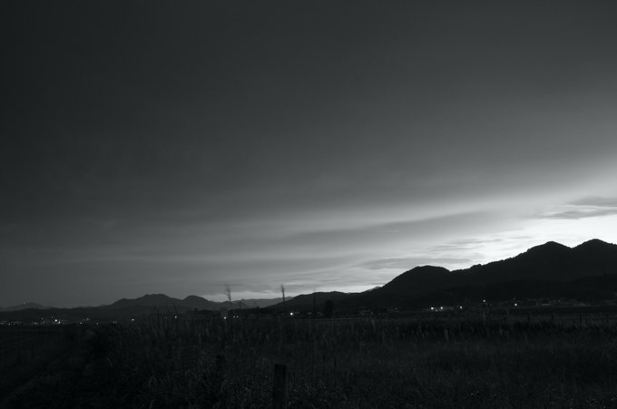 Black and white photograph of a rural landscape at dusk or dawn, showing silhouetted mountains and grass against a cloudy sky.