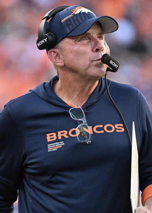 A close-up of a coach wearing a navy blue "BRONCOS" hoodie and matching cap, sporting a Sony coaching headset, standing on the sidelines of a football game.