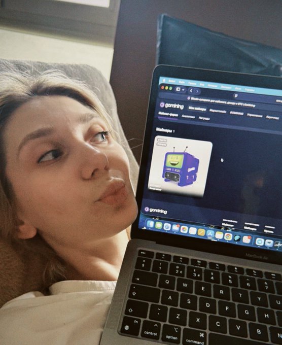 A young woman lies down next to an open MacBook Air, looking at the screen which displays a website called 'gambimg' with an image of a blue TV-headed character.