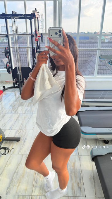 A woman taking a mirror selfie in a gym, wearing a white t-shirt and black shorts, with a treadmill and weight equipment visible in the background.