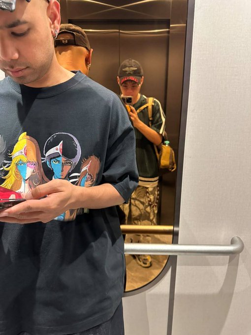 Two people are visible in what appears to be an elevator; one in the foreground wears an anime-themed t-shirt and looks at a phone, while another in the background takes a mirror selfie wearing a cap and holding a beverage.