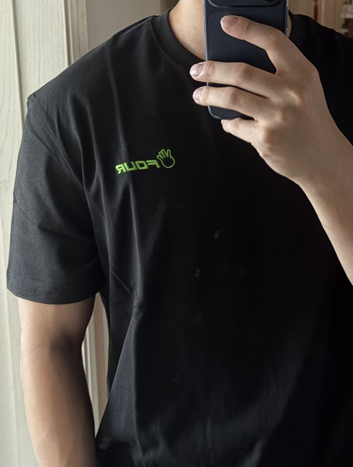 A person takes a mirror selfie wearing a black t-shirt featuring the green "fourdot" logo with a hand icon, promoting merchandise for the Fourmeme community.