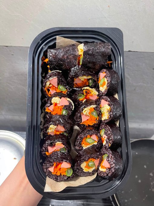A black plastic takeout container holds neatly arranged slices of gimbap (Korean seaweed rice rolls) filled with vegetables, egg, and ham, wrapped in dark purple-black rice.