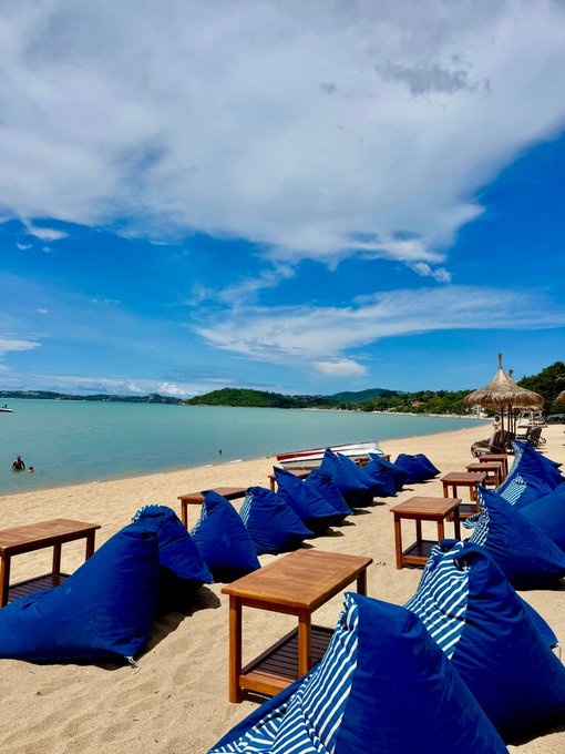 A scenic beach view featuring rows of blue bean bag chairs and wooden tables on the sand, with turquoise water and a cloudy blue sky in the background.