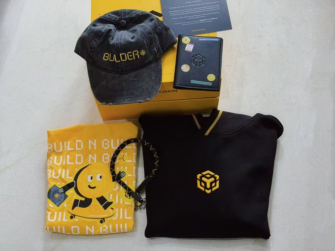 The image features a collection of items related to the brand "Build N Build" displayed on a table or countertop. These items include a black hoodie, a yellow shirt with a cartoon character, a hat, a wallet, and a box containing a book and a card. The text in the image reads: "Received BNB feeding during Pizza Day Thanks Sandy @0xsandy1913 Those working hard to build BNB, Sandy can see it all All have a chance to receive peripheral feeding".