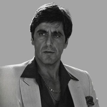 A black-and-white photo of a man in a white blazer over a black shirt, wearing a gold chain, resembling a classic Hollywood portrait of a 1970s actor.