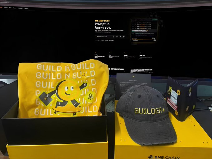 BNB Chain branded merchandise including a yellow 'BUILD' t-shirt with a smiley mascot, a black 'BUILDER' cap, and trading cards, displayed in front of a large curved screen showing 'BNB AGENT STUDIO'.