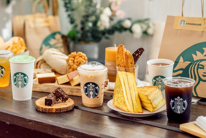 An arrangement of Starbucks coffee cups, iced drinks, cakes, pastries, and paper bags on a wooden table.