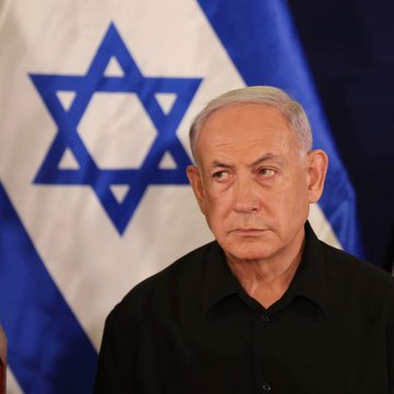 A serious-looking man with gray hair wearing a black shirt stands in front of an Israeli flag featuring the blue Star of David.