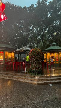 A rainy outdoor scene of a restaurant patio or market stall area, featuring red counters, green canopies, and red lanterns hanging overhead.