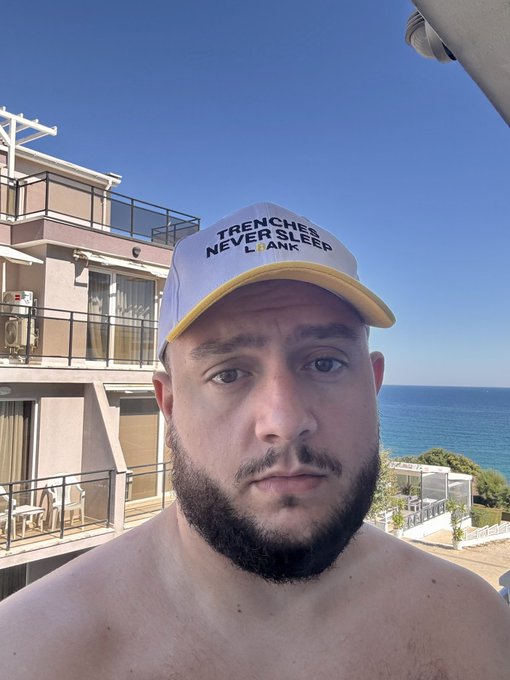 A selfie of a bearded man wearing a white and yellow trucker hat that reads 'TRENCHES NEVER SLEEP L BANK,' taken on a balcony with a clear blue sky and ocean in the background.