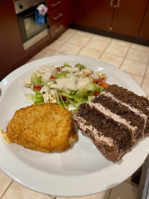A white plate holds a meal consisting of a breaded fried patty, a slice of layered chocolate cake, and a green salad topped with creamy dressing and croutons.