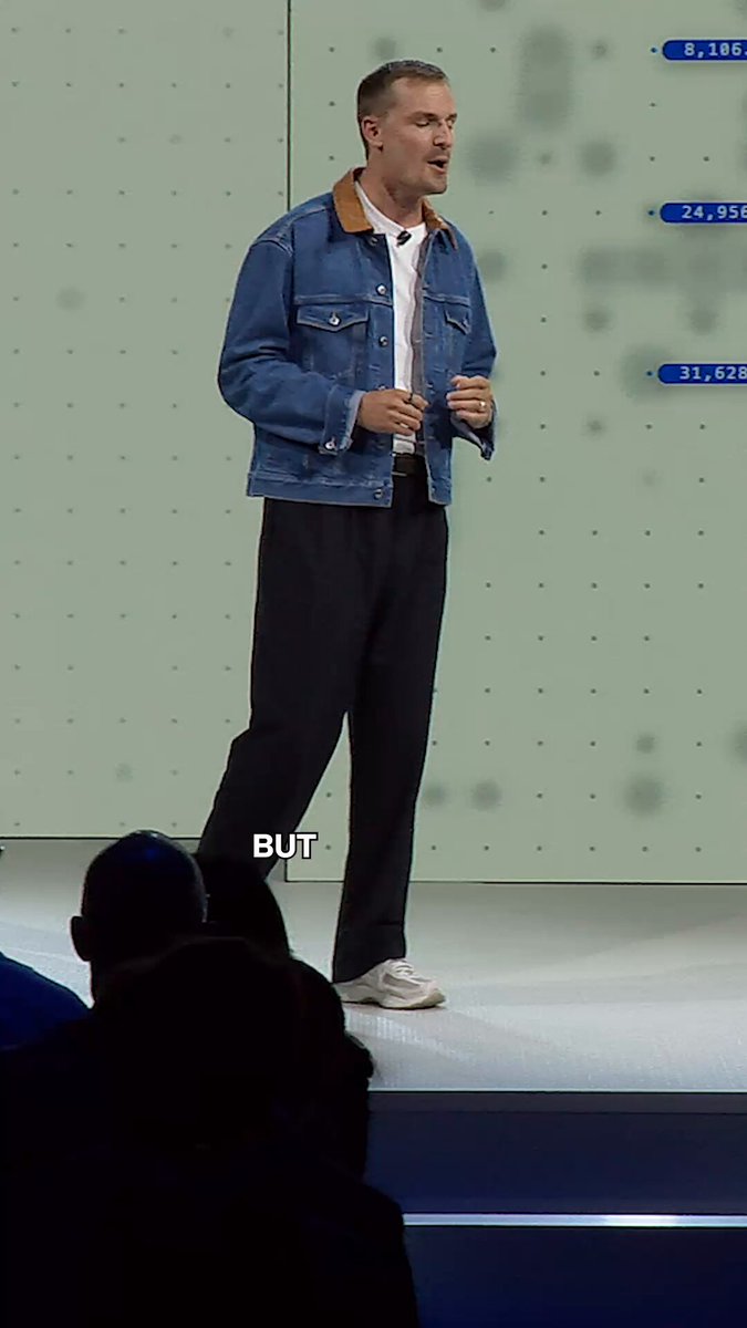 The image shows a man standing on stage with his hands up, possibly giving a speech or presentation. He is wearing a blue jacket and black pants. In front of him, there are several people sitting in chairs, indicating that this might be an event where multiple speakers are present. The background features a green wall with white dots scattered across it, which could be part of the stage setup or decorations for the event.