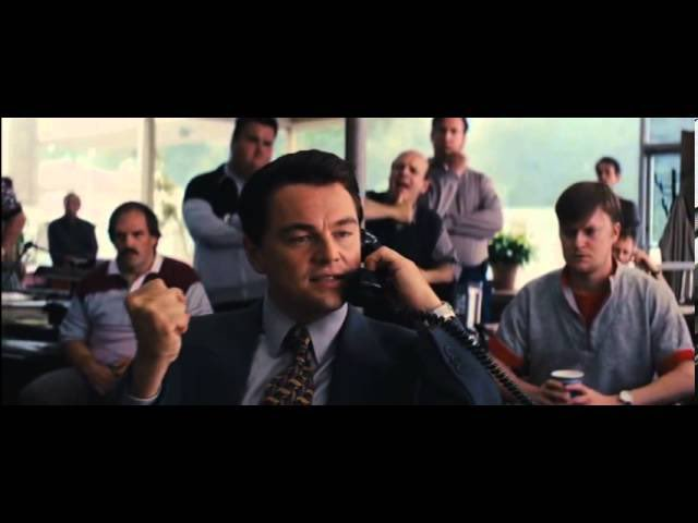 A film still featuring Leonardo DiCaprio in a suit on a telephone, clenching his fist in a triumphant gesture with onlookers behind him.