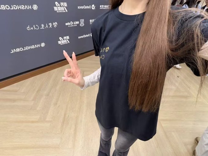 A young woman with long dark hair poses wearing a black t-shirt with gold text, throwing up a peace sign in front of a step-and-repeat backdrop featuring logos for DataCrypto, HYGH, and UNTX.