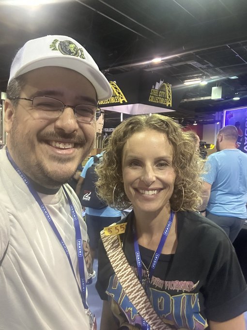 A man and woman pose for a photo at what appears to be an expo, both wearing 'AMAZON' lanyards; the man's cap has a lion logo, and a 'STEEL CITY COLLECTIBLES' banner is visible in the background.