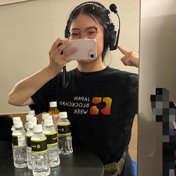 A woman wearing glasses takes a mirror selfie while pointing out that her headset is worn backwards, with "BLOCKCHAIN WEEK JAPAN" visible on her black t-shirt and a cluster of water bottles on the table in front of her. The caption "まって！！！ これ左右反対につけとった！" ("Wait!!! I had this on the wrong way!") humorously matches her gesture of pointing at the incorrectly worn headset.