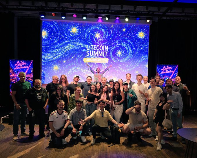 The image shows a group of people posing for a photo at a Litecoin Summit event, with a large screen behind them displaying a starry night sky theme. The attendees are standing close together, smiling and enjoying themselves as they capture this memorable moment.