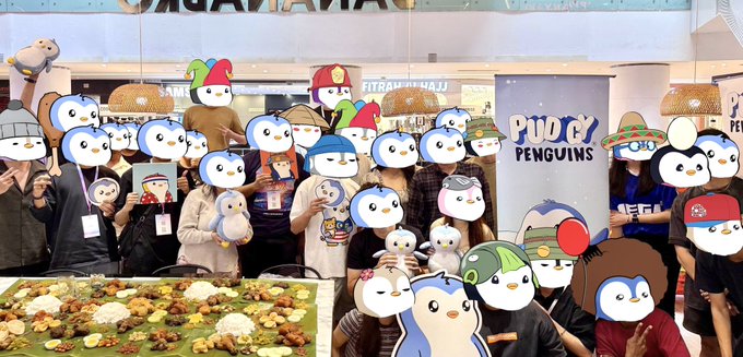 A group photo of people wearing Pudgy Penguins plush penguin masks and hats at what appears to be a Pudgy Penguins community event, with various costumed characters posing together.