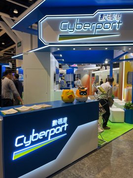 A trade show booth for "Cyberport" (數碼港) features mascots and branding, with people gathered around promotional materials on the counter. The booth appears to be related to the "$Floki" partnership mentioned, showcasing a collaboration display.