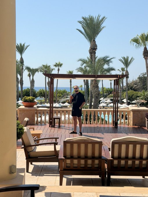A man in sunglasses and black attire enjoys a sunny "vibe check" on a luxurious resort terrace overlooking palm trees and the ocean, framed by elegant wooden pergolas and cushioned seating.