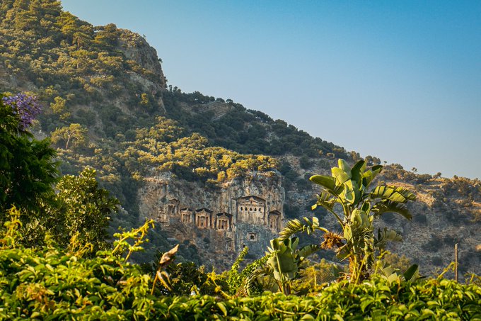 Ancient Lycian rock-cut tombs carved into a mountainous cliff face, surrounded by lush green vegetation and banana plants under a clear blue sky.