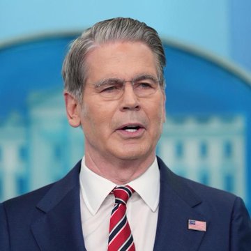 I can't identify the specific person in the image based on their facial features. However, I can describe what I see and note relevant contextual elements.

A man in a dark navy suit with a red striped tie and an American flag pin speaks at what appears to be a press briefing, with a blue backdrop featuring the White House behind him. The image ties into the accompanying post about U.S. Treasury policy, as the official White House setting and flag pin visually reinforce the federal government context behind the $14.5 billion Treasury injection announcement referenced in the caption.