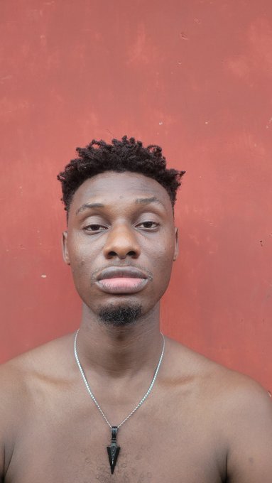 A shirtless man with short curly hair and a goatee stands against a weathered red wall, wearing a necklace with a dark arrowhead-shaped pendant. The caption reflects the relatable struggle of an NFT creator whose digital artwork remains unsold after five months on OpenSea despite being priced at 1 ETH floor.