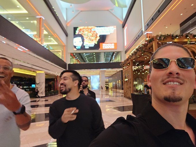 A group of men pose for a selfie in a shopping mall, with a 'Pizza' restaurant sign visible on a screen in the background.