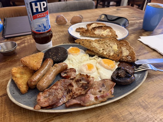 A hearty breakfast plate featuring sausages, eggs, bacon, black pudding, hash browns, mushrooms, and toast, served with a bottle of HP Sauce and a blue coffee mug.