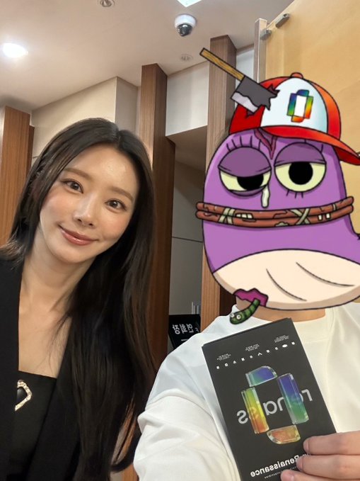 A selfie-style photo of a smiling Asian woman with long dark hair standing next to a purple cartoon character sticker wearing a cap, with a Microsoft logo visible on a product box held in the foreground.