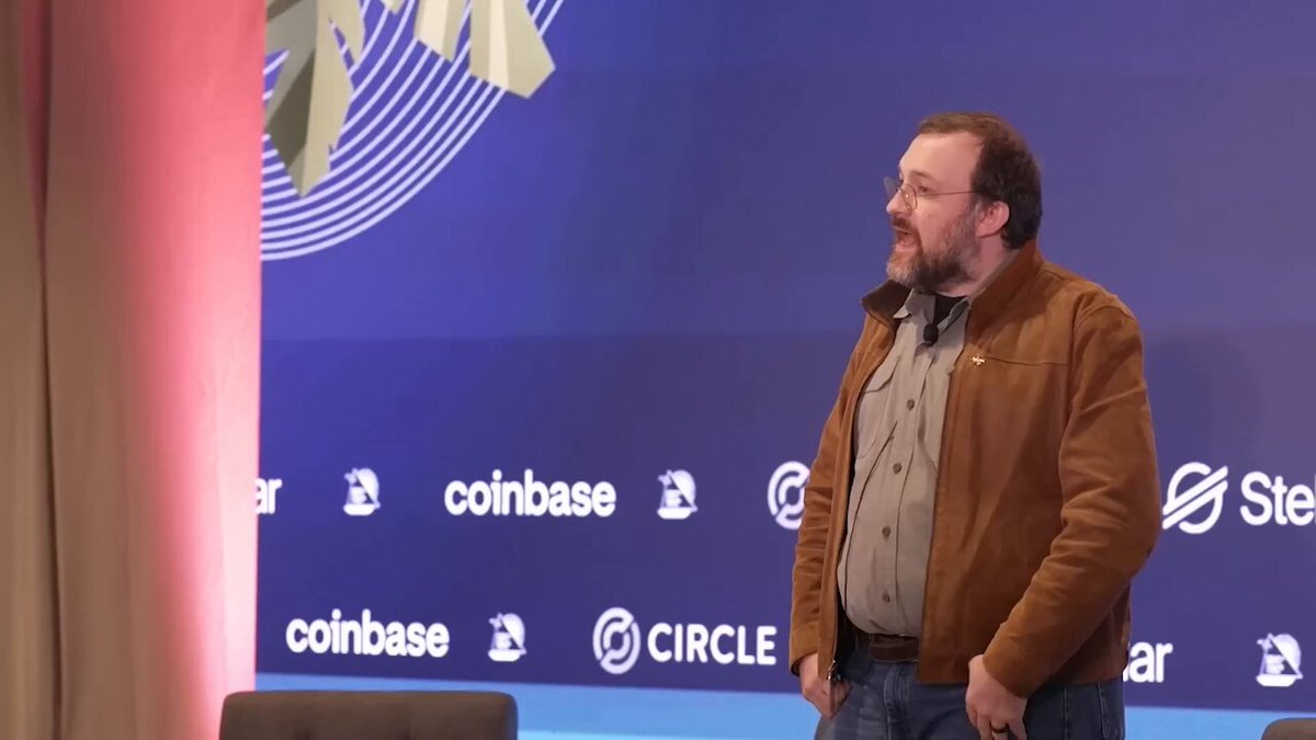 The image features an older man wearing glasses standing in front of a blue wall adorned with white logos for Coinbase and Circle. The man appears to be giving a speech or presentation at an event, possibly related to cryptocurrency or blockchain technology.