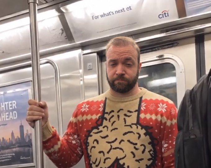 A bearded man with blonde hair wearing a festive red sweater featuring a yellow snowman design, standing inside a subway car and holding onto a metal pole with a Citi advertisement visible behind him.