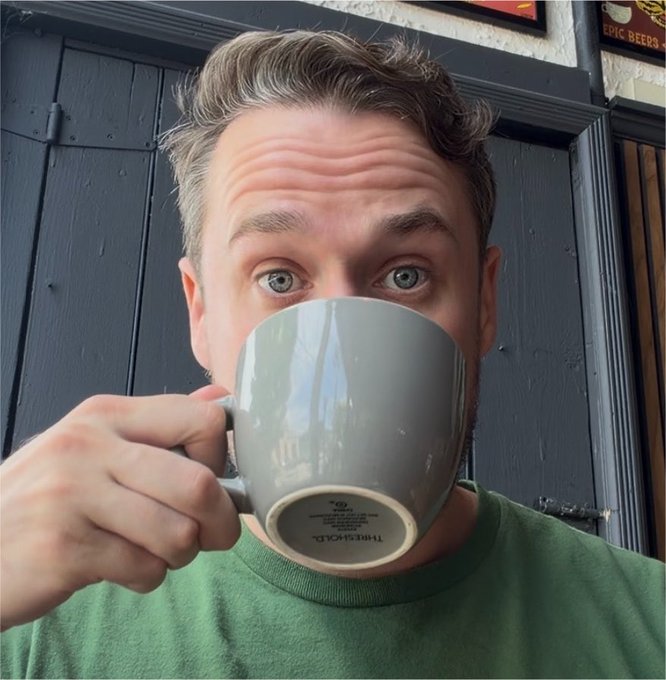 A close-up selfie of a man with light brown hair and blue eyes drinking from a grey 'THRESHOLD' branded mug, wearing a green sweater, with dark wooden doors and an 'EPIC BEERS' sign in the background.