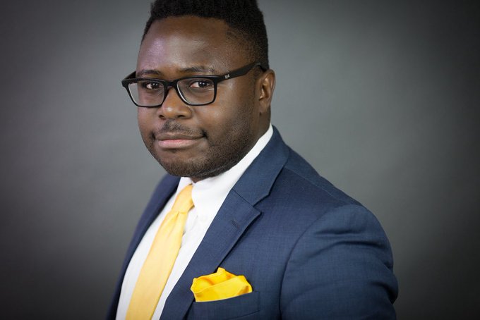 A professional headshot of a Black man wearing glasses, a navy blue suit, white shirt, and yellow tie with a matching pocket square.